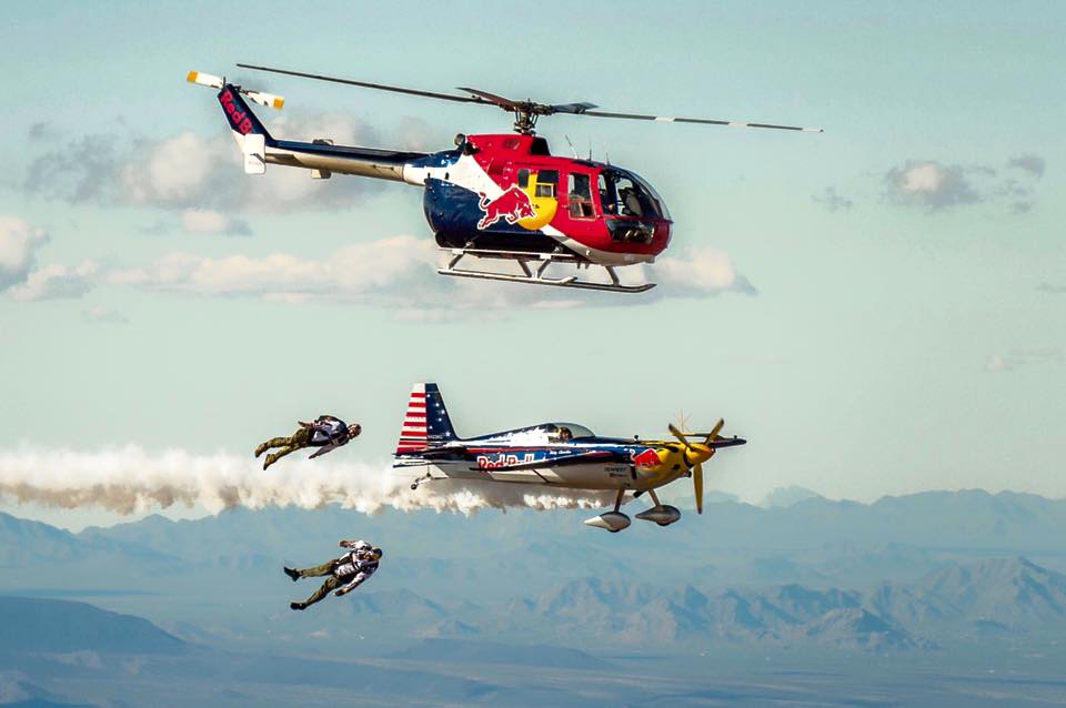 Luke Aikins flies in a wingsuit with a plane and helicopter in the background. 