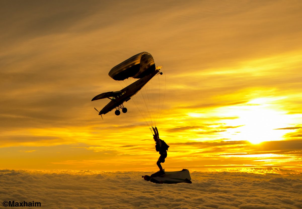 Pete flying under canopy at sunset while seemingly standing on a wingsuit skydiver