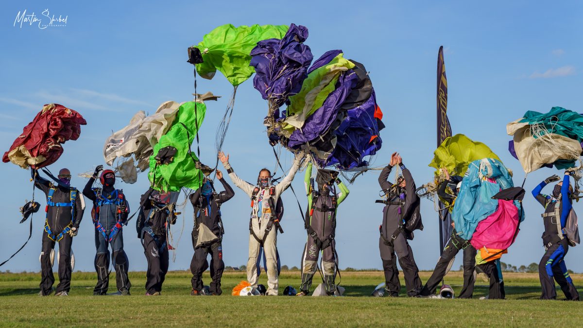 10 skydivers stand in the landing area and throw their canopies in the air for a photo op